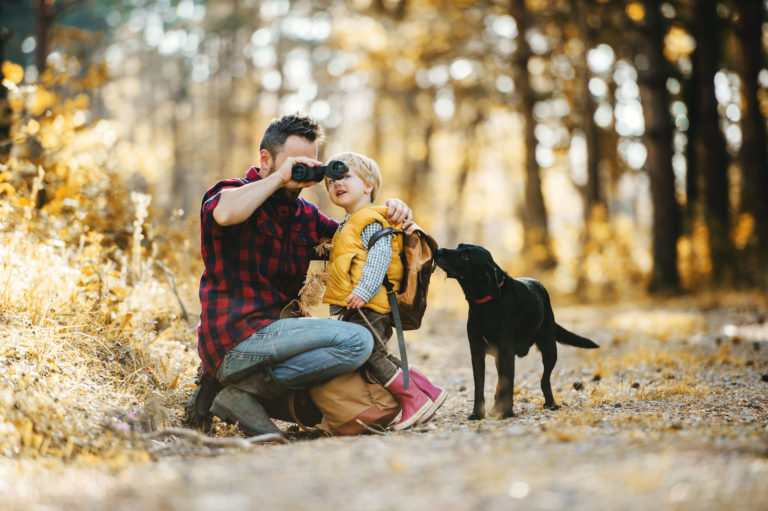 Vater mit Kind und Hund stehen im Wald und schauen durch ein Fernglas